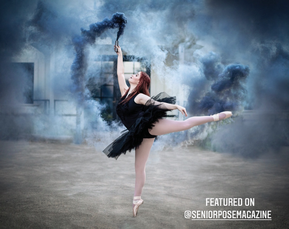 teenage girl with long red hair and black tutu in an arabesque pose holding a blue smoke bomb