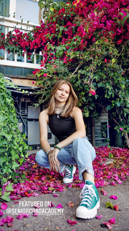 teenage girl in black top and jeans and green conversesquatting with pink flowers around her 