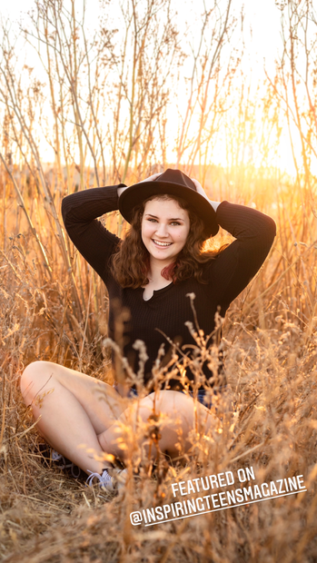teenage girl with brown hair wearing all black and a black hat sitting a wheat field at sunset