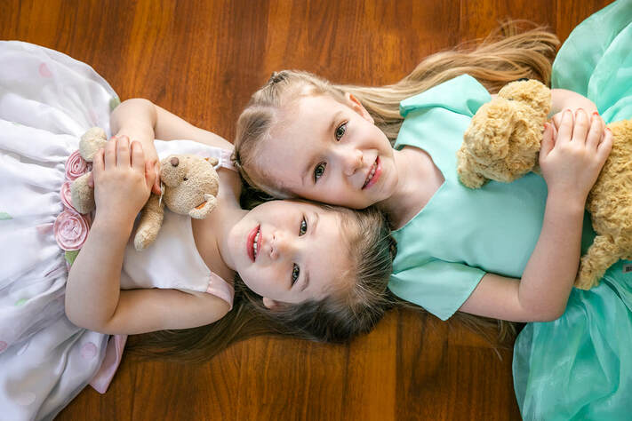 young girls laying on floor with heads together holding stuff animals