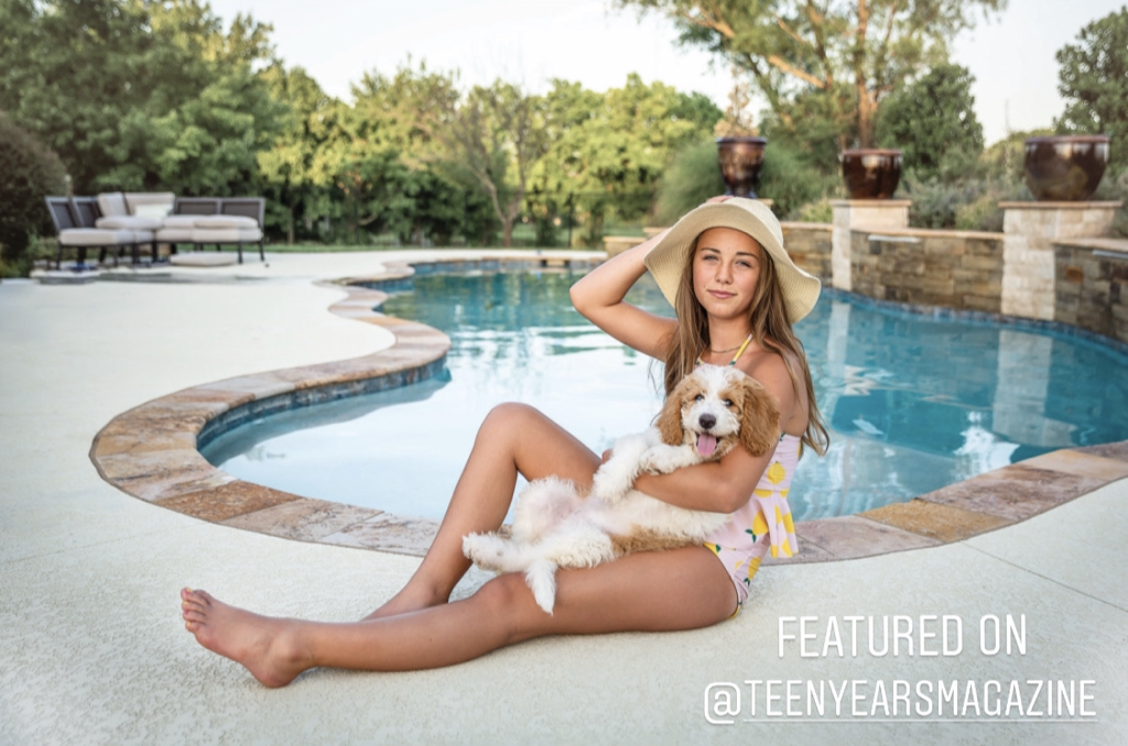 teenage girl with blond hair, wearing a hat and bathing suit, holding her dog, by a swimming pool